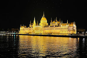 Das beleuchtete ungarische Parlamentsgebäude bei Nacht. Im Vordergrund ist die Donau zu sehen. 