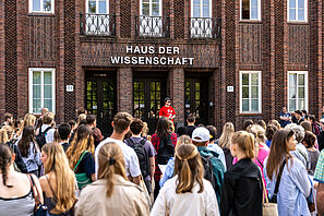 Eine Menschenmenge steht vor einem Gebäude, an dem Haus der Wissenschaft steht. Im Hintergrund sieht man einen Menschen in einem roten TU T-Shirt, der ein Blatt Papier in der Hand hält und der Menschenmenge etwas erzählt