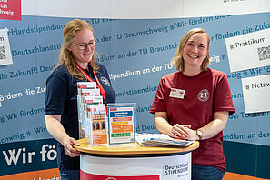 Zwei Personen stehen an einem Messestand. Auf der Wand im Hintergrund steht "Deutschlandstipendium an der TU Braunschweig". Die Personen lachen.