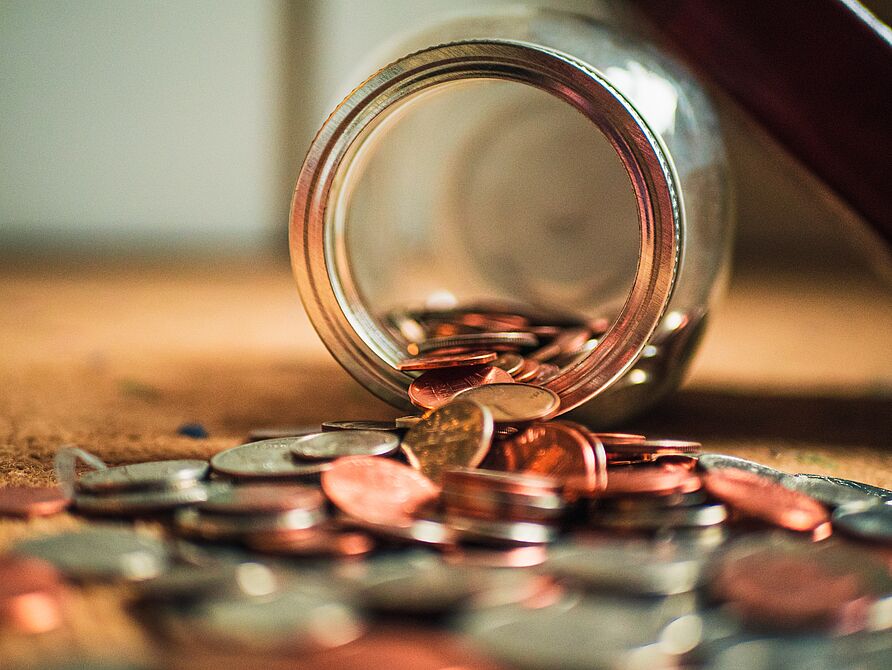 Ein auf der Seite liegendes Glasgefäß, aus dem Münzen heraus auf einen Tisch gefallen sind.