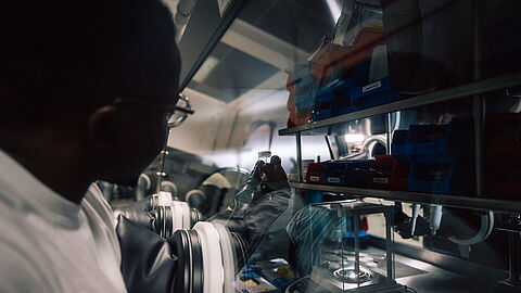 A student in a lab coat, wearing protective gloves that are permanently attached, works with samples behind glass