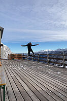 Die Studentin springt auf der Terasse einer Berghütte in die Luft. Im Hintergrund ist das Alpenpanorama Österreichs zu erkennen. 