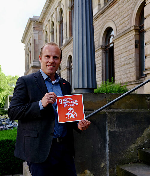 Dr.-Ing. Mark Mennenga steht vor dem Altgebäude der TU Braunschweig und hält das Schild für das 9. SDG.