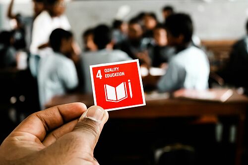 A hand holds a small red-and-white card bearing the words “Quality Education.”