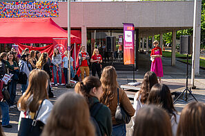 Eine Gruppe Menschen steht um ein Podium herum. Auf dem Podium steht eine Frau in einem pinken Kleid und hält eine Rede. Neben ihr hängt eine Flagge auf der HIT und herzlich willkommen steht. Im Hintergrund ist ein roter Pavillon zu sehen und ein Gebäude mit einem Banner auf dem steht #buntstadt Zeit für Demokratie - gegen Hass und Hetze