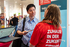Zwei Personen im Gespräch, eine trägt ein rotes T-Shirt mit der Aufschrift 'ZUHAUSE IN DER ZUKUNFT', im Hintergrund ein Banner der Universität Braunschweig