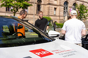 Im Vordergrund ist die Motorhaube und Windschutzscheibe eines Autos. Auf der Motorhaube steht "Peter L. Reichertz Institut für medizinische Informatik". Im Hintergrund sind drei Personen zu erkennen