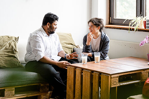 Zwei Studierende sitzen auf einem Sofa in einem Universitätscafé und unterhalten sich. Auf dem Tisch stehen zwei Gläser mit Kaffee.