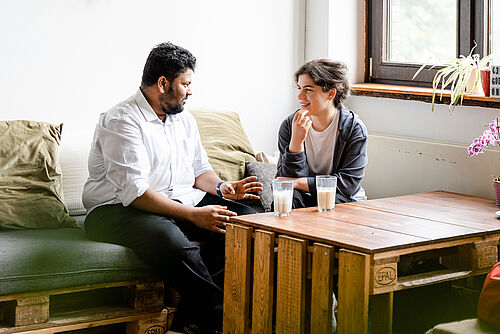 Two students are sitting on a sofa in a university café and chatting. There are two glasses of coffee on the table.