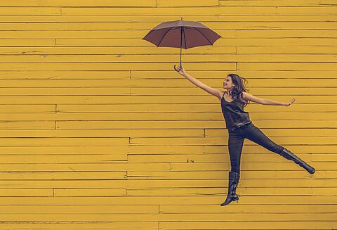 Frau mit Schirm fliegt scheinbar vor gelber Wand